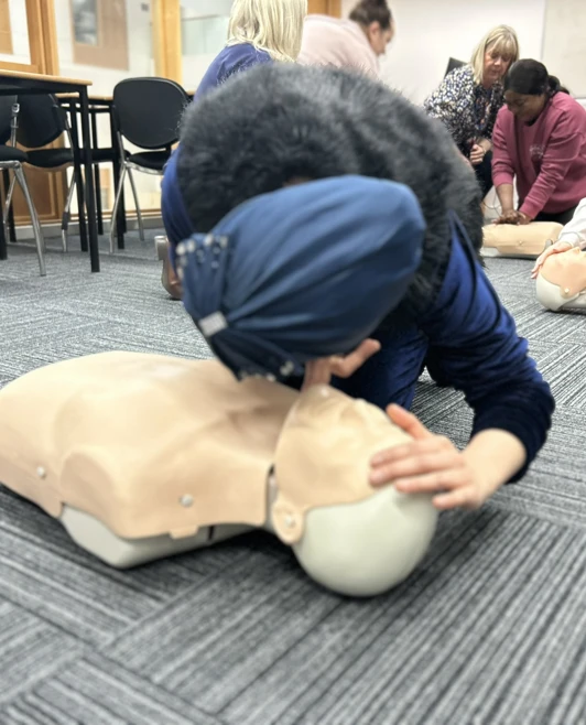 A woman in a blue headscarf is performing mouth-to-mouth resuscitation on a CPR mannequin as part of a first aid training session, with other participants practicing in the background. A woman in a blue headscarf is performing mouth-to-mouth resuscitation on a CPR mannequin as part of a first aid training session, with other participants practicing in the background.